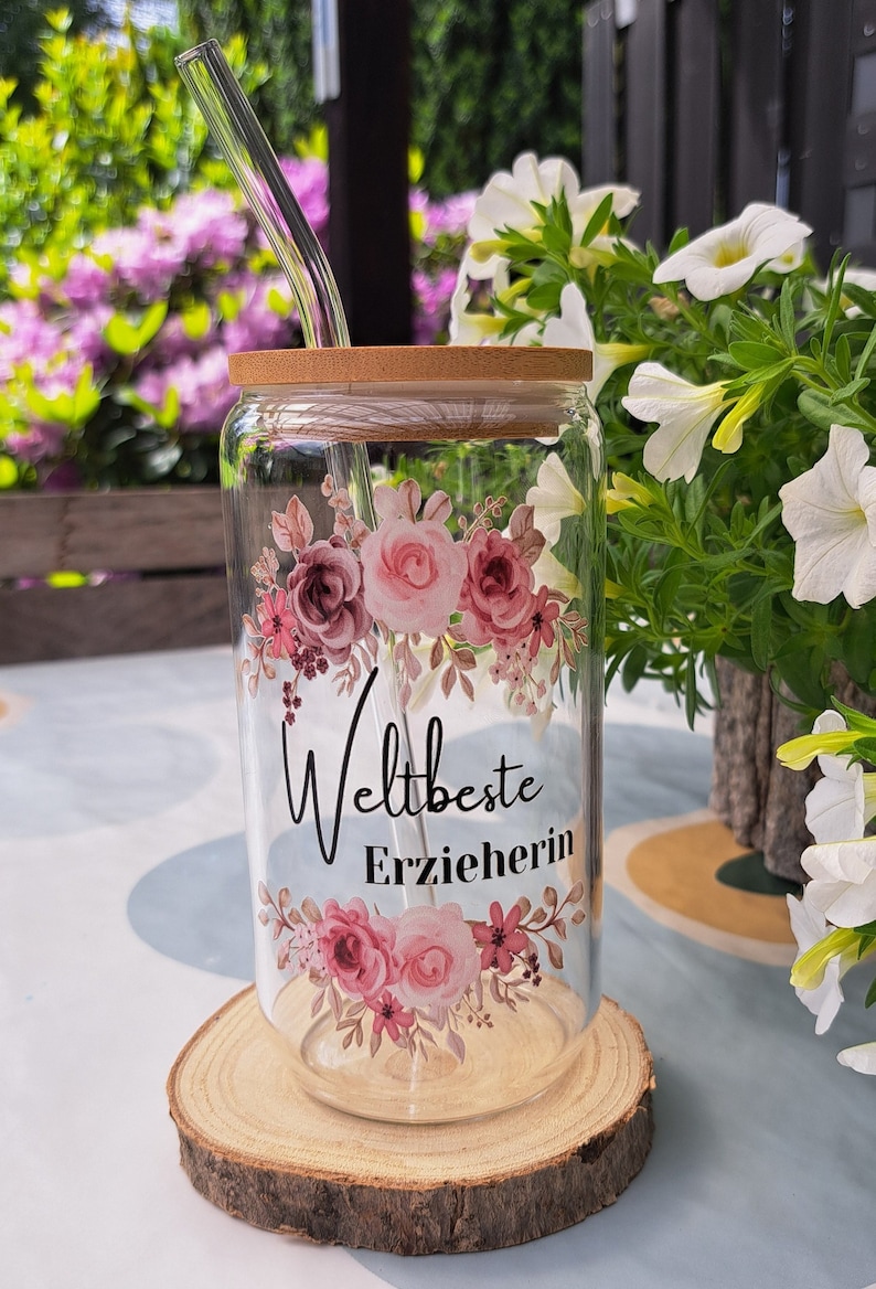 Op de afbeelding: Een doorzichtig glazen beker met een bamboe deksel en een glazen rietje, voorzien van een bloemmotief van roze en mauve rozen en de zwarte tekst "Weltbeste Erzieherin" op de voorzijde, geplaatst op een ronde houten schijf.