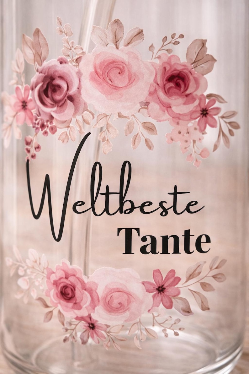 Może przedstawiać: Przezroczysta szklanka z kwiatowym wzorem przedstawiającym r&oacute;żowe r&oacute;że i liście. Tekst "Weltbeste Tante" jest nadrukowany czarną czcionką, łączącą pismo odręczne z bezszeryfową, pomiędzy kwiatowymi motywami na gładkiej, cylindrycznej powierzchni.