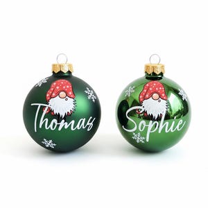 K&ouml;nnte beinhalten: Zwei gr&uuml;ne Weihnachtskugeln mit goldenen Kappen. Jede Kugel zeigt ein Zwergen-Design mit roter M&uuml;tze und wei&szlig;em Bart sowie einen Namen in wei&szlig;er Schrift: "Thomas" und "Sophie". Schneeflocken-Akzente sind ebenfalls vorhanden.