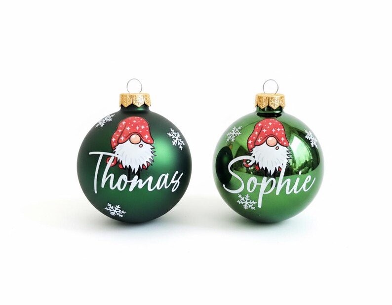 K&ouml;nnte beinhalten: Zwei gr&uuml;ne Weihnachtskugeln mit goldenen Kappen. Jede Kugel zeigt ein Zwergen-Design mit roter M&uuml;tze und wei&szlig;em Bart sowie einen Namen in wei&szlig;er Schrift: "Thomas" und "Sophie". Schneeflocken-Akzente sind ebenfalls vorhanden.