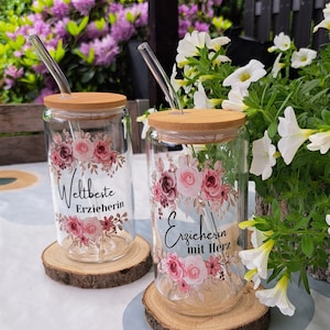 Könnte beinhalten: Zwei Glasbecher mit Blumendesigns und Holzdeckeln. Die Becher haben Strohhalme und stehen auf Holz-Untersetzern. Der Text auf den Bechern lautet "Weltbeste Erzieherin" und "Erzieherin mit Herz".