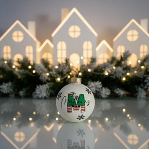 以下が含まれることがあります： 「Matthias」という名前と緑と赤の「M」の文字が入った白いクリスマスオーナメント。オーナメントは金色のキャップと雪の結晶で飾られています。背景には光る白い家があります。