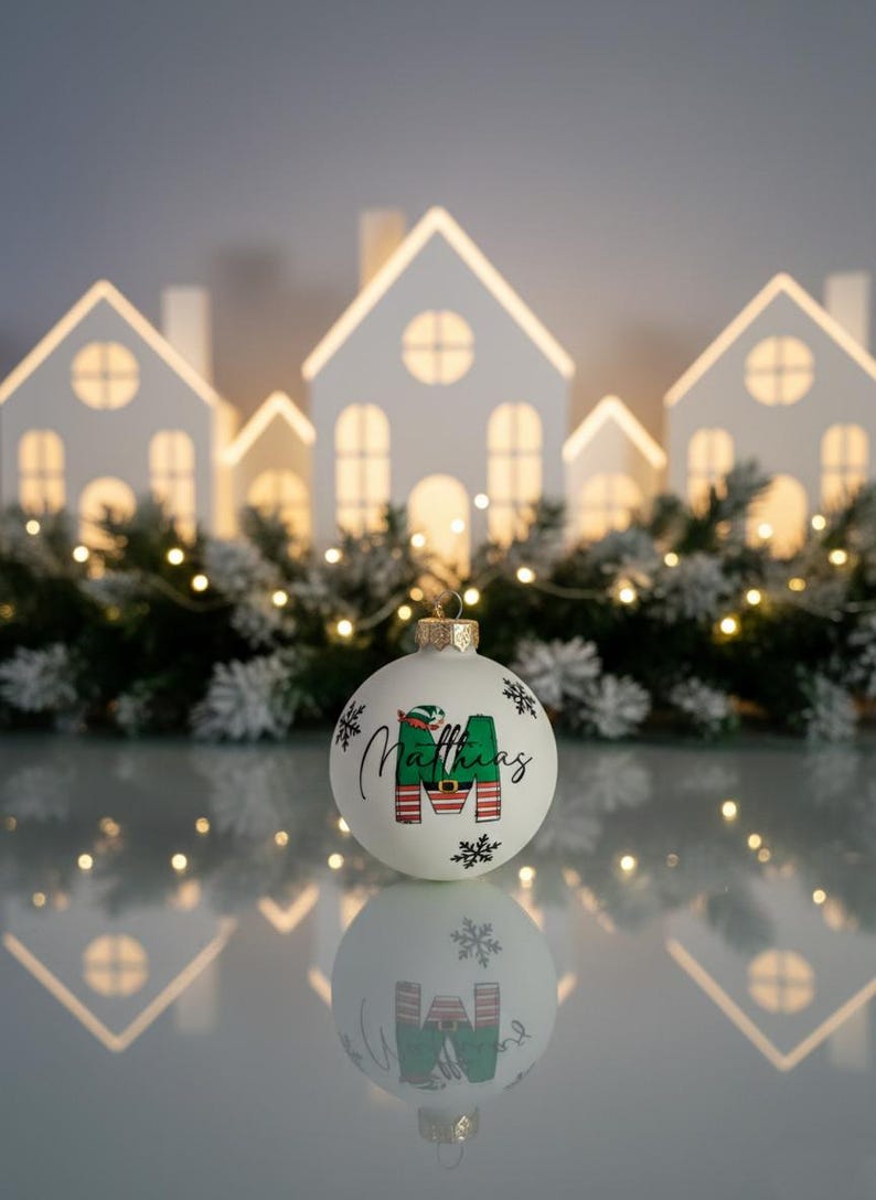 以下が含まれることがあります： 「Matthias」という名前と緑と赤の「M」の文字が入った白いクリスマスオーナメント。オーナメントは金色のキャップと雪の結晶で飾られています。背景には光る白い家があります。