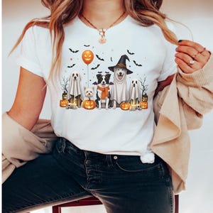 Könnte beinhalten: Weißes T-Shirt mit einem Halloween-Motiv. Das Design zeigt Cartoon-Hunde in Kostümen, Kürbisse, Fledermäuse und einen orangefarbenen Ballon. Die Hunde tragen einen Hexenhut, ein Geisterkostüm und einen orangefarbenen Schal. Das T-Shirt hat einen klassischen Rundhalsausschnitt.