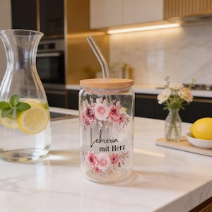 K&ouml;nnte beinhalten: Ein klares Trinkglas mit einem Bambusdeckel und einem gebogenen Glasstrohhalm. Das Glas ist mit einem Blumenmuster in Rosa- und Gr&uuml;nt&ouml;nen verziert, wobei der schwarze Schriftzug "Lehrerin mit Herz" mittig zwischen den Blumen angebracht ist.