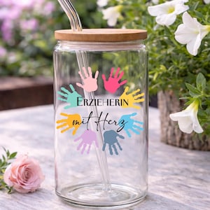 May include: A clear glass tumbler with a wooden lid and a glass straw. The tumbler is decorated with colourful handprints and the text "ERZIEHERIN mit Herz". The background features flowers.
