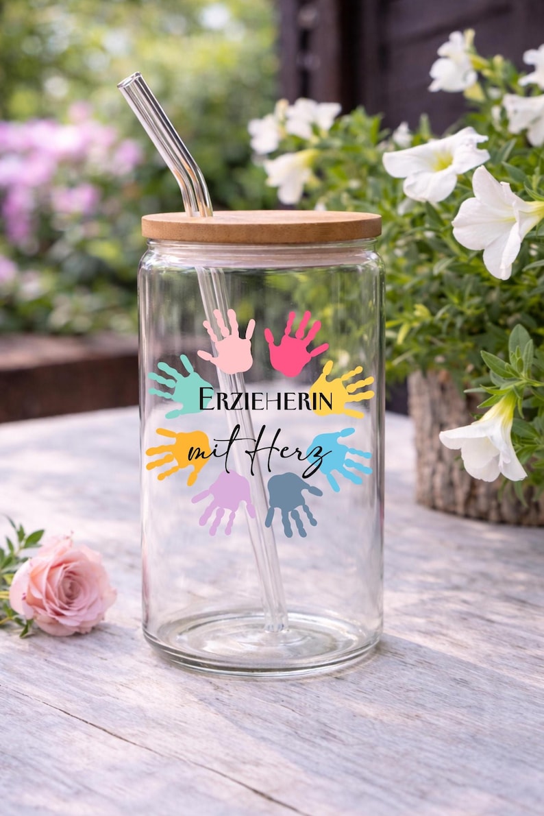 May include: A clear glass tumbler with a wooden lid and a glass straw. The tumbler is decorated with colourful handprints and the text "ERZIEHERIN mit Herz". The background features flowers.