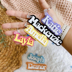 May include: Five colourful acrylic keychains with names written in white. The names are Katie, Mackenzie, Amelia, Layla, Joseph, and Eleanor.