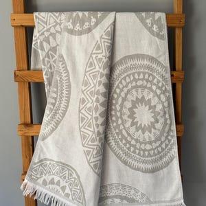 Peut inclure: Une serviette à motifs gris clair et blancs avec un motif géométrique est drapée sur une échelle en bois. La serviette présente de grands motifs circulaires avec des formes complexes et un bord à franges. La serviette est en matière douce.