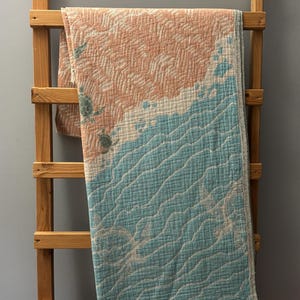 Peut inclure: Une couverture aux couleurs pêche, blanche et turquoise, drapée sur une échelle en bois. La couverture présente un motif de plage avec un motif texturé et une bordure à franges. Dimensions : environ 150 cm x 200 cm.