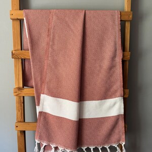 Könnte beinhalten: Ein terrakottafarbenes Handtuch mit einem geometrischen Rautenmuster und einem breiten weißen Streifen. Das Handtuch wird auf einem hölzernen Leiterregal präsentiert und hat weiße Fransen am unteren Rand. Das Handtuch besteht aus einem weichen, saugfähigen Material.