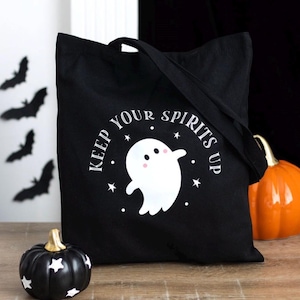 Könnte beinhalten: Eine schwarze Tragetasche mit einer weißen Grafik eines lächelnden Geistes mit rosa Wangen, umgeben von kleinen Sternen und dem Text "KEEP YOUR SPIRITS UP" in einer weißen Serifenschrift. Die Tasche hat zwei Tragegriffe und eine rechteckige Form.