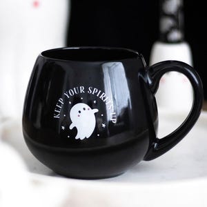Könnte beinhalten: Schwarze Keramik-Tasse mit runder Form und großem Henkel. Die Tasse zeigt eine weiße Geistergrafik mit dem Text "KEEP YOUR SPIRITS UP" in einem kreisförmigen Design. Die Tasse steht auf einer weißen Oberfläche.