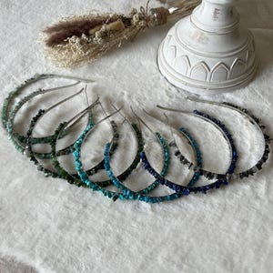Peut inclure: Plusieurs bandeaux pour cheveux ornés de pierres précieuses dans des tons de vert, de bleu et de gris. Chaque bandeau est composé d'une bande en métal argenté ornée de petites pierres de forme irrégulière. Ils sont disposés sur une surface blanche texturée.