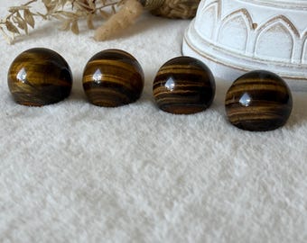 May include: Four polished tiger's eye gemstones, each shaped as a smooth, rounded dome. These stones feature distinct, alternating bands of golden-brown and dark brown colors with a reflective, chatoyant luster across their curved surfaces.
