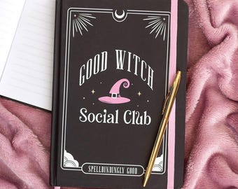 May include: A black hardcover journal with a pink elastic band closure, featuring white text that reads Good Witch Social Club and Spellbindingly Good, a pink witch hat graphic, and a gold-toned ballpoint pen resting on the cover.