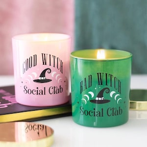 May include: Two glass candles with lit wicks. The pink candle features black text reading Good Witch Social Club with a witch hat icon. The green candle features black text reading Bad Witch Social Club with a witch hat icon. Both have gold lids.
