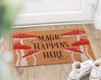 May include: A rectangular doormat with a brown background and red and white mushroom illustrations. The text "MAGIC HAPPENS HERE" is printed in brown. White sneakers with black star patterns are next to the mat.