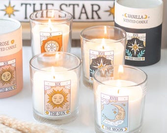May include: Four lit candles in clear glass jars, each featuring a tarot card label: The Sun XIX, The Moon XVIII, The Star XVII, and The Wheel of Fortune X. Two cylindrical cardboard tubes are present, labeled Vanilla Scented Candle and Rose Scented Candle.