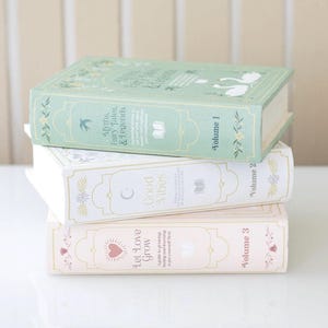 Op de afbeelding: Een stapel van drie rechthoekige decoratieve boeken met pastelkleurige covers. Het bovenste boek is saliegroen met witte zwaanillustraties en de tekst Myths, Fairy Tales & Legends Volume 1. Het middelste boek is wit met de tekst Good Vibes Volume 2. Het onderste boek is lichtroze met de tekst Let Love Grow Volume 3.