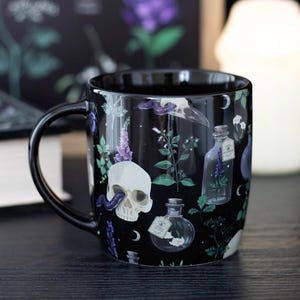 Op de afbeelding: Een zwarte keramische mok met een herhalend patroon van witte menselijke schedels, paarse slangen, glazen apothekersflessen met etiketten en diverse groene botanische planten, versierd met kleine halve maan iconen over het oppervlak.