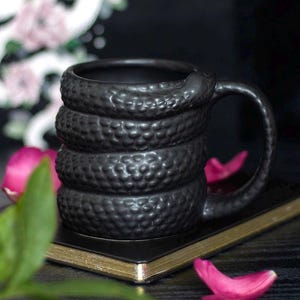 Puede incluir: Una taza de cerámica negra mate con un diseño tridimensional de serpiente enroscada alrededor del exterior. La superficie de la taza tiene una apariencia texturizada y escamosa, y el asa está integrada en la forma del cuerpo de la serpiente.