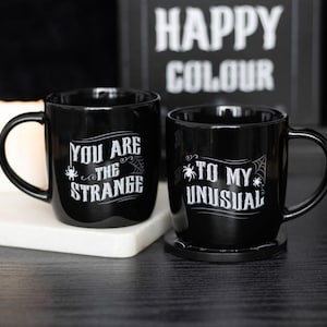 Könnte beinhalten: Zwei schwarze Keramikbecher mit weißem Text. Auf einem Becher steht "YOU ARE THE STRANGE" mit einer Spinnen- und Netzgrafik. Der andere Becher sagt "TO MY UNUSUAL" mit einer Spinne und einem Netz. Ein Schild im Hintergrund zeigt "HAPPY COLOUR".