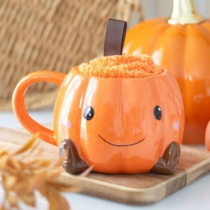 May include: An orange ceramic pumpkin-shaped mug with a handle, black eyes, and a smiling face. The mug is filled with orange material and has a brown ribbon attached. A larger pumpkin is in the background. The mug is on a wooden board.