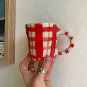 May include: A ceramic mug with a red and white check design. The handle features red and white stripes. The mug is held in a hand, with a plain white wall in the background. A framed picture is visible.