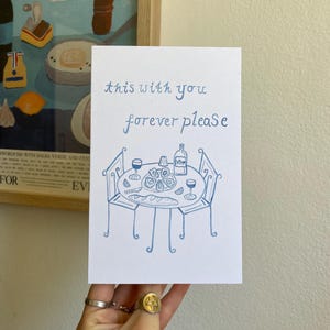 May include: A white card with blue handwritten text that reads "this with you forever please." Below the text is a blue illustration of a table set for two with wine, oysters, and bread. A hand holds the card.