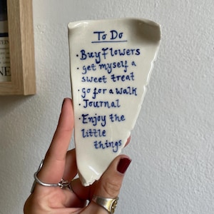 May include: A white ceramic trinket dish shaped like a jagged piece of paper with blue handwritten text that reads: To Do, Buy flowers, get myself a sweet treat, go for a walk, Journal, Enjoy the little things. The dish has a smooth, glossy finish.