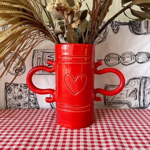 May include: A bright red ceramic vase featuring a cylindrical body with two large, curved side handles. The front of the vase is embossed with a centred heart shape and horizontal line details. The vase is filled with dried, light brown foliage.