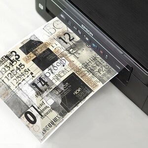 May include: A black and silver printer with a sheet of paper coming out of the paper tray. The paper has a collage of black and white text and numbers.