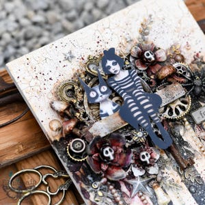 May include: A mixed media art piece with a white background. The piece features a black and white illustration of a girl with a cat. The girl is wearing a striped dress and has a black bow in her hair. The piece is decorated with various embellishments, including gears, flowers, and other decorative elements.
