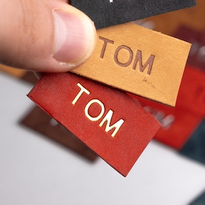 Peut inclure: Gros plan sur plusieurs étiquettes en cuir avec le mot "TOM" en relief dans différentes couleurs, notamment noir, marron, rouge et vert. Les étiquettes sont disposées en quinconce.