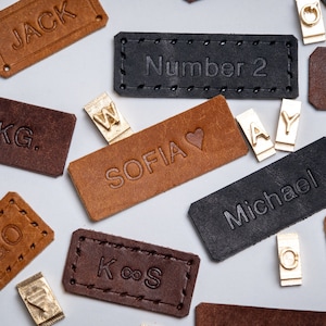 Op de afbeelding: Een assortiment van bruine, zwarte en donkerbruine leren labels met verschillende namen en woorden erop gegraveerd, waaronder "Jack", "Sofia", "Michael", "Nummer 2", "K&S", "E&O", ".KG.", en "GeoF".