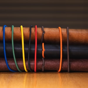 May include: A stack of three leather journals in shades of brown and black, secured with colorful elastic bands. The journals have a rustic, textured appearance, with visible stitching and a minimalist design. The elastic bands are blue, green, yellow, red, and orange.