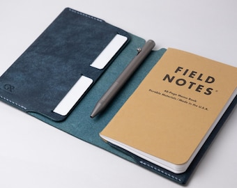 May include: A blue leather notebook cover with a pen and a Field Notes memo book inside. The cover has a pocket for cards and a stitched edge. The memo book is brown with black text that reads "FIELD NOTES 48-Page Memo Book Durable Materials/Made in the U.S.A."