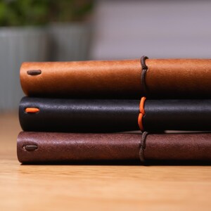 May include: A stack of three leather-bound journals in shades of brown and black. The journals are closed, with a visible orange accent on the black journal. The journals are bound with a brown cord.