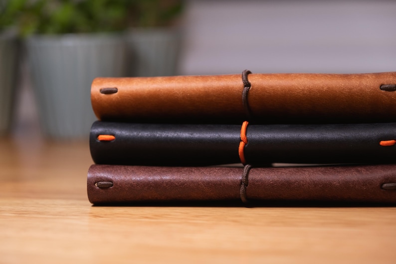May include: A stack of three leather-bound journals in shades of brown and black. The journals are closed, with a visible orange accent on the black journal. The journals are bound with a brown cord.