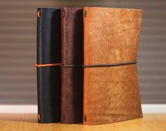May include: Three leather-bound journals with elastic closures. The journals are brown, black, and a light brown color. The journals are sitting on a wooden surface.