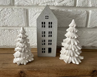 May include: Three white ceramic Christmas tree ornaments and a gray concrete house ornament. The ornaments are on a wooden surface.