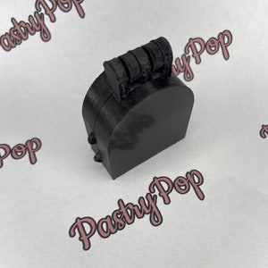 Puede incluir: Una caja negra impresa en 3D, con bisagras, con una base curva y una serie de crestas en la parte superior. La caja está sobre una superficie blanca con las palabras "Pasty Pop" en escritura rosa.