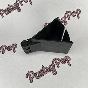 Puede incluir: Un soporte para teléfono de plástico negro con un diseño triangular. El soporte está diseñado para sostener un teléfono en posición vertical.
