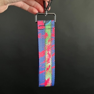 Pode incluir: Um cordão colorido com um fecho de metal. O cordão apresenta um fundo azul com um padrão vibrante de respingos de tinta vermelha e verde. O fecho de metal é prateado e preso a uma peça de metal retangular.