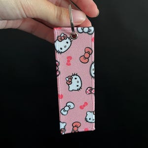 May include: Pink rectangular keychain featuring a repeating pattern of Hello Kitty faces, bows, and pink bone shapes. The keychain has a silver metal ring for attaching keys. The background is black.