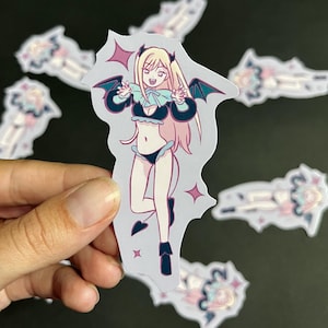 Puede incluir: Una pegatina con un personaje de estilo anime con cabello rubio, alas de murciélago negras y un atuendo negro y azul. El personaje está sonriendo y rodeado de estrellas. Otras pegatinas son visibles en el fondo.
