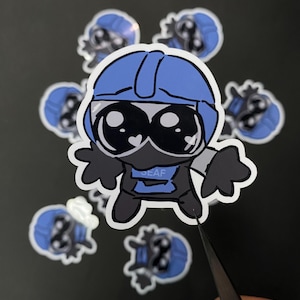 Könnte beinhalten: Eine Sammlung von Aufklebern mit einer Cartoonfigur mit blauem Helm, schwarzer Gesichtsbedeckung und schwarzem Outfit. Die Figur hat große Augen und ein herzförmiges Detail. Das Wort "GEAF" ist auf dem Outfit zu sehen. Die Aufkleber befinden sich auf einer dunklen Oberfläche.