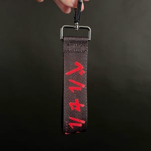 Puede incluir: Correa de llavero negra y roja con caracteres japoneses. La correa está sujeta a un cierre metálico de color plateado. El texto japonés es en negrita, rojo. El fondo es negro liso.