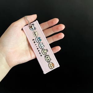 Könnte beinhalten: Ein rosa rechteckiger Schlüsselanhänger mit einem silbernen Schlüsselring. Der Schlüsselanhänger zeigt gestickte Cartoon-Figuren und das Wort "CHIKAWA" in Weiß. Die Charaktere sind in verschiedenen Farben und Designs.